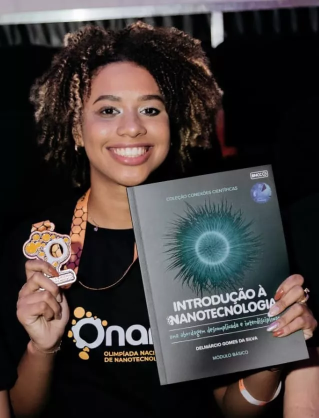 Discente Michele com a medalha numa mão e o livro introdução à nanotecnologia na outra. Ela sorri ao posar pra câmera. 